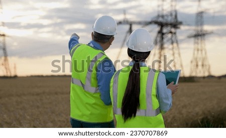 Electricians with tablet walk along rural field against blurry power lines