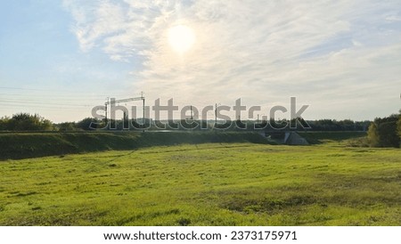 Electrified railroad tracks are laid along the mound among grassy meadows and mixed forests. A metal bridge on a concrete base was built across the creek. Sunny autumn weather and blue sky with clouds