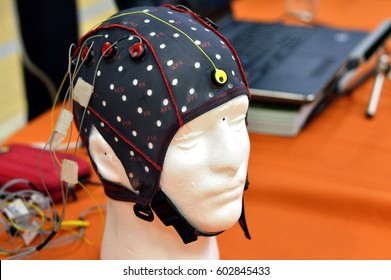 The electroencephalogram (EEG) head cap with flat metal discs (electrodes) attached to a white plastic model’s head shown in a science exhibition, with laptops blurred at the background.