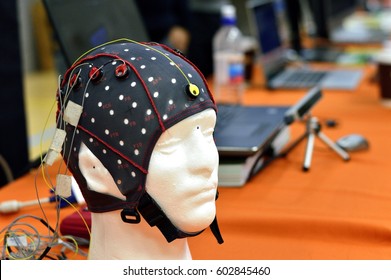 The electroencephalogram (EEG) head cap with flat metal discs (electrodes) attached to a white plastic model’s head shown in a science exhibition, with laptops blurred at the background.