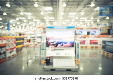 Electronic Department Store With Bokeh Blurred Background. Television Retail Shop, TVs Display On Shelf At Wholesale Store. Defocused Warehouse Interior Technology Aisle, Shelves And Shopping Cart.