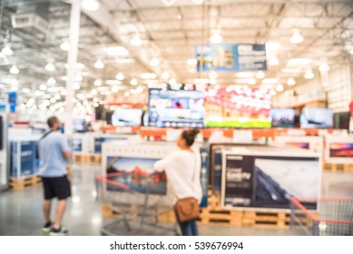 Electronic Department Store With Bokeh Blurred Background. Television Retail Shop, TVs Display On Shelf At Wholesale Store. Defocused Warehouse Interior Technology Aisle, Shelves And Customer Shopping