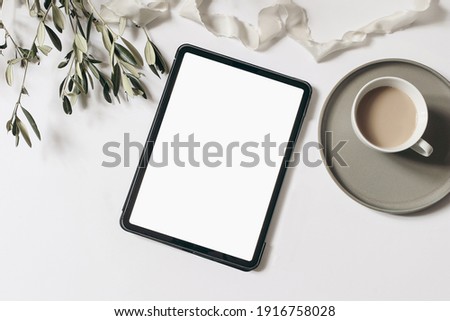 Electronic device mock-up. Closeup of tablet with empty screen. Cup of coffee, olive tree branches and ribbon isolated on white table background. Flat lay, top view. Blogger design. Home office.
