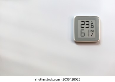 Electronic Indoor Gray Plastic Digital Thermometer On White Wall Showing Temperature, Humidity Level In House, Smiling Smiley Face Indoors In Daylight Sun And Parallel Stripes Of Shadow