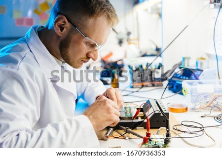 Electronic laboratory worker connects cirquit board with wires and clamps for testing and metering. Man in white robe and protective glasses working at desk. Blue and white color scheme.