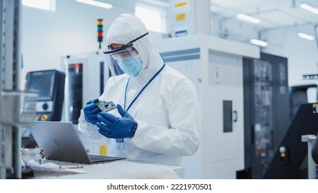Electronic Manufacturing Factory: Engineer in Sterile Coverall Working on Laptop Computer, Examining a Circuit Board with Microchips and Testing New Electrical Equipment.