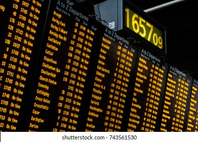 An electronic passenger information board inside a busy train station giving passengers information about destinations, departures, arrivals, delays, timings and platform numbers with a digital clock.