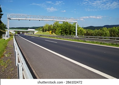 Electronic Toll Gate Over A Highway In A Wooded Landscape. White Trucks, Bridge And Forested Mountains In The Background. White Clouds In The Blue Sky.