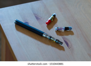 An Electronic Vape Pen, With CBD And THC Cartridges Around It On A Wooden Table.