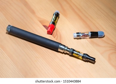 An Electronic Vape Pen, With CBD And THC Cartridges Around It On A Wooden Table.