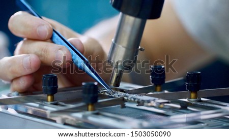 Electronics engineer repairing chip in workshop, hands closeup. Repairman heating chipset using soldering blow dryer and taking hot melting element forceps. Repair and maintenance service center.