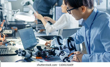 Electronics Engineer Works With Robot, Manipulating Its Responses And Checking Data With Laptop Computer. Computer Science Research Laboratory With Specialists Working.