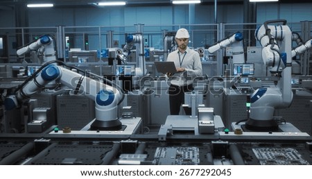 Electronics Factory Manager Working on Laptop at an Industrial Manufacture with Robotic Arms. Specialist Monitoring Data on a Computer, Checking and Planning Production Process