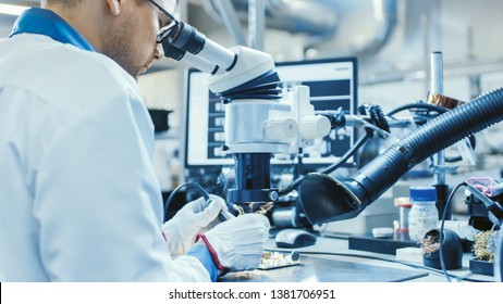 Electronics Factory Worker In White Work Coat Is Soldering A Printed Circuit Board Through A Digital Microscope. High Tech Factory Facility.