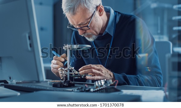 Electronics\
Maintenance Engineer Soldering Motherboard, Microchip and Circuit\
Board, Looking through Magnifying Glass, Consults Personal\
Computer. Electronics Repair and\
Testing