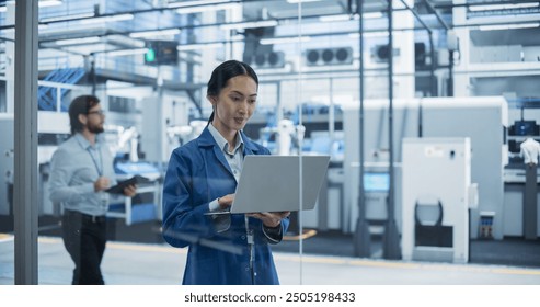 Electronics Manufacturing Technician in a Blue Coat Working on a Laptop Computer at an Assembly Plant. Asian Female Specialist Developing, Maintaining, Setting Up Software for Automated Production - Powered by Shutterstock