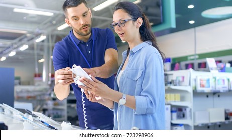 In The Electronics Store Consultant Gives Professional Advice To A Young Woman, She Considers Buying New Tablet Computer And Needs Expert Opinion. Store Is Modern, Bright And Has All The New Devices.