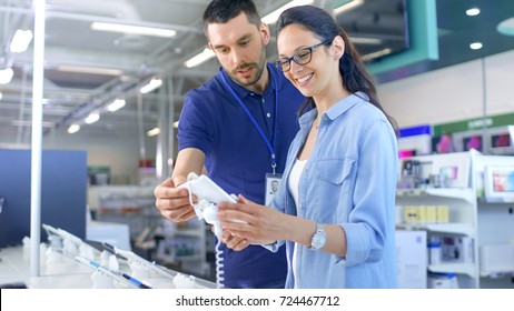 In The Electronics Store Consultant Gives Professional Advice To A Young Woman, She Considers Buying New Tablet Computer And Needs Expert Opinion. Store Is Modern, Bright And Has All The New Devices.