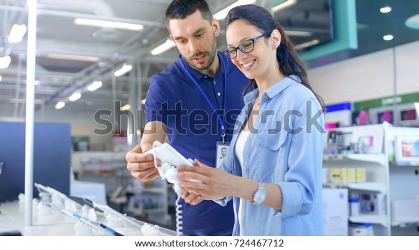 In the Electronics Store Consultant Gives\
Professional Advice to a Young Woman, She Considers Buying New\
Tablet Computer and Needs Expert Opinion. Store is Modern, Bright\
and Has all the New Devices.