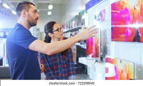 In The Electronics Store Professional Consultant Provides Expert Advice To A Young Woman Who Looks For A  New TV To Buy. In This Bright, Modern Store All The Latest TV Models Are Hanging On The Wall.