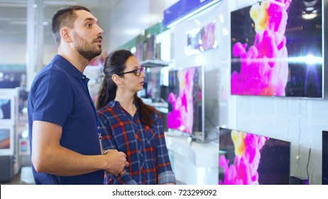 In The Electronics Store Professional Consultant Provides Expert Advice To A Young Woman Who Looks For A  New TV To Buy. In This Bright, Modern Store All The Latest TV Models Are Hanging On The Wall.