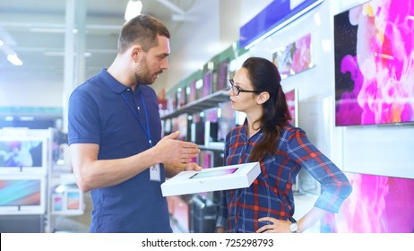 In The Electronics Store Professional Consultant Provides Expert Advice On Tablet Computer Specifications For Beautiful Young Woman. Store Is Bright, Modern And Has All The Latest Devices.