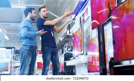 In The Electronics Store Professional Consultant Shows TV's To A Young Man, They Talk About Specifications And What Model Is Best For His Home. Store Is Bright, Modern And Has All The Latest Models.