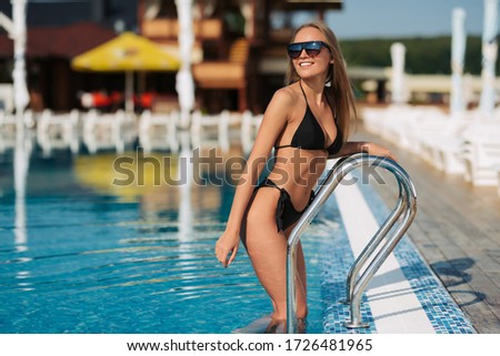 Elegan woman in the black bikini on the sun-tanned slim and shapely body is posing near the swimming pool, wearing hat and stylish sunglasses