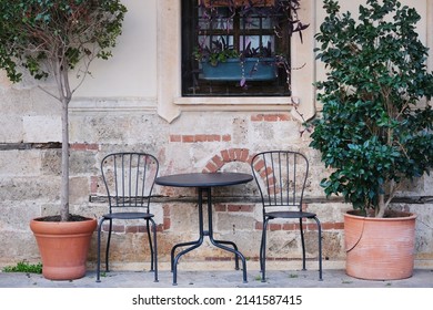 An Elegance Metal Table And Empty Black Chairs Next To Potted Trees  In A Street Cafe  Against The Background Of A Window And A Wall Of An Old Stone Building 