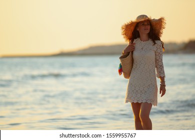 Elegant 40 Year Old Woman In White Dress And Straw Hat On The Ocean Coast At Sunset Walking.