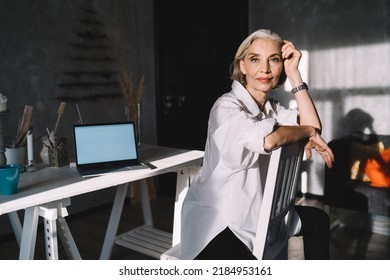 Elegant 50 Years Old Caucasian Businesswoman Sitting On Chair And Looking At Camera. Concept Of Small Business And Entrepreneurship. Modern Successful Woman. Dark Art Studio With Sunny Lighting