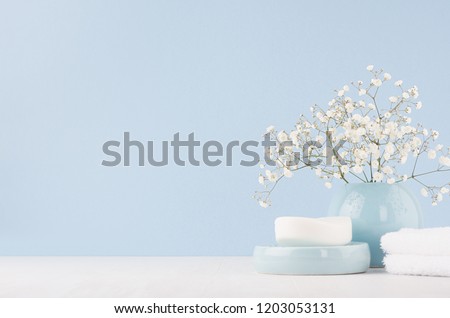 Elegant acessories for dressing table - soft pastel blue ceramic bowls, white flowers, products for skin and body care on white wood board and blue wall.