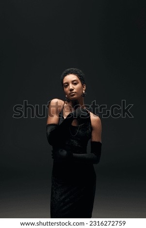 Elegant african american woman in gloves, golden necklaces and little dress looking at camera while standing isolated on black, high fashion and evening look