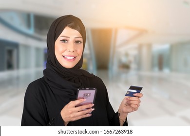 Elegant Arab Woman In Abaya Standing At Mall Holding Mobile Phone And Plastic Card. Businesswoman With Stylish Hijab Paying With Credit Card While Shopping Online Using Mobile.