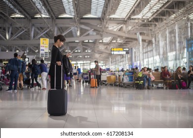 Elegant Asian Businesswoman Checking E-mail On Mobile Phone With Suitcase In Airport And Airport Terminal Blurred Crowd Of Travelling People On The Background. 