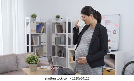 Elegant Asian Chinese Pregnant Ceo Woman Standing In The Office Is Drinking Tea And Touching Her Tummy While Reviewing Project On Laptop