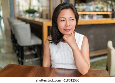 Elegant Asian middle-aged businesswoman pensive portrait. Beautiful mature Chinese business woman relaxing in restaurant bar urban stylish living,