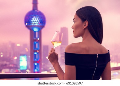 Elegant Asian Woman In Gown Drinking White Wine Glass At Rooftop Bar Terrace Looking At City Lights Skyline View Of Shanghai In Sunset. Luxury Travel Or High End Lifestyle.