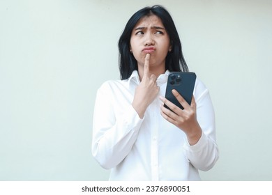 elegant asian young business woman daydreaming using smartphone wearing white formal shirt with finger on chin looking up confused what to buy in online shopping sale standing over isolated background - Powered by Shutterstock