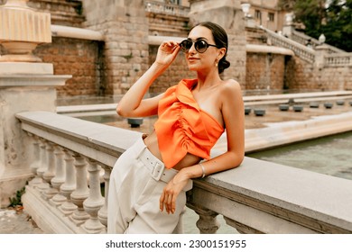 Elegant attractive stylish european woman with dark hair wearing orange top and white pants is golding glasses and looking straight with lovely smile outdoor in the city  - Powered by Shutterstock