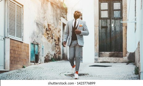 An Elegant Bald Bearded African Man In A Fashion Costume With Tie And Orange Socks Is Walking Down The Antique Street Of A European City; A Fancy Black Guy In A Suit Is Leaving The Door Of His House