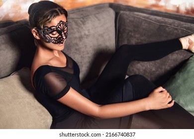 Elegant ballerina in a masquarade mask wearing a black dress sits on the Sofa