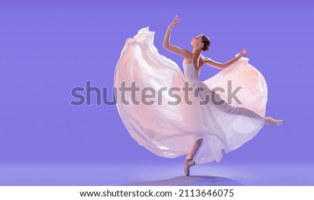 elegant ballerina in pointe shoes is dancing in a long flying white skirt on a lilac light background