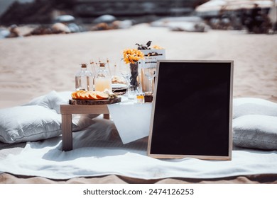 Elegant beach picnic setup with a wooden table featuring sliced citrus fruits, glass bottles, and a bouquet of yellow flowers. Blank chalkboard for text. Background includes sand and beach umbrellas - Powered by Shutterstock