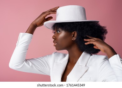Elegant beautiful African American woman wearing classic white hat, blazer, posing in studio, on pink background. Close up profile portrait. 