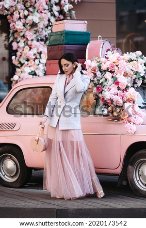 Elegant beautiful female fashion model in luxury tulle dress and jacket with long brunette hair pose at pink retro car with flower decoration outdoors. Professional makeup and stylish outfit concept