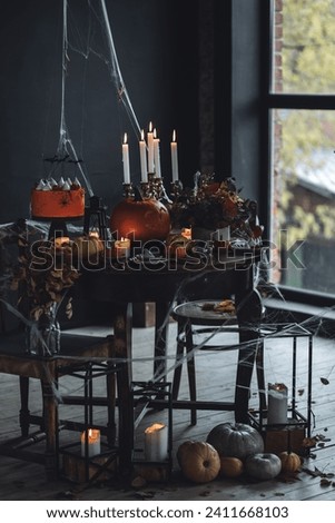 Elegant beautiful Halloween party table decor. Burning candled in vintage chandelier, handmade cake with meringue ghosts, handmade jack-o-lantern. Dark background, wooden old furniture, spider web