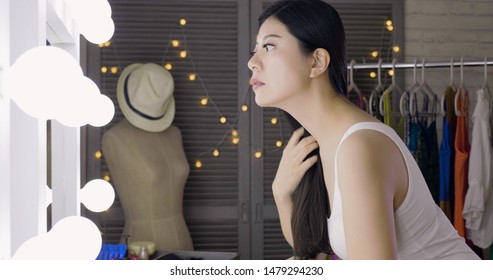 Elegant Beautiful Young Asian Woman Touching Her Long Black Hair Looking In Mirror In Dressing Room Backstage. Stylish Haircare Concept In Makeup Room. Female Actress Before Changing Clothes By Rack.