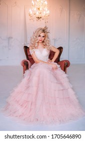 elegant beautiful young princess girl. Blonde queen woman sitting in old medieval vintage armchair. Golden crown pink vintage long dress. Backdrop shines crystal chandelier white interior classic