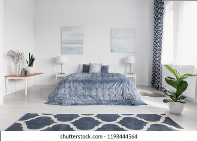 Elegant bedroom interior with big comfortable bed with blue bedding, paintings on the wall and patterned carpet on the floor, real photo